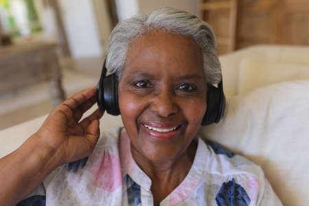 Portrait of senior african american woman sitting on sofa wearing headphones smiling. retreat, retirement and happy senior lifestyle concept.の写真素材