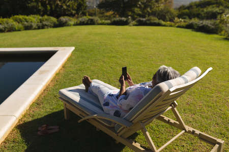 Senior african american woman using smartphone in deckchair by swimming pool in sunny garden. retreat, retirement and happy senior lifestyle concept.の写真素材
