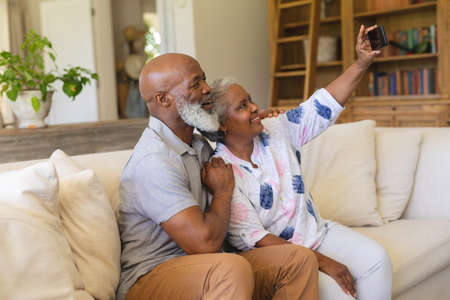 Senior african american couple sitting on sofa using smartphone taking selfies. retreat, retirement and happy senior lifestyle concept.の写真素材