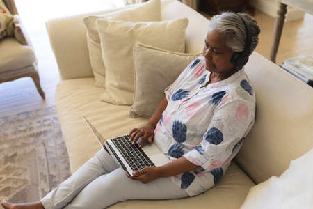 Senior african american woman sitting on sofa wearing headphones using laptop. retreat, retirement...