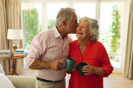 Senior caucasian couple kissing on cheek smiling and holding mugs in living room. retreat,...