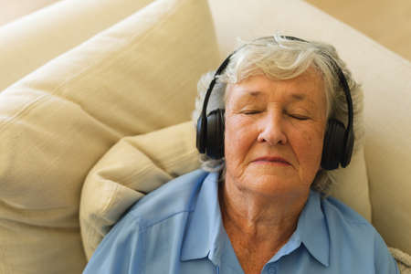 Senior caucasian woman sitting on sofa wearing headphones with eyes closed. retreat, retirement and happy senior lifestyle concept.の写真素材