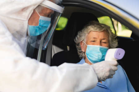 Medical worker wearing ppe suit taking temperature of senior caucasian woman sitting in car....