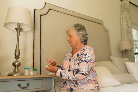 Senior caucasian woman sitting on bed holding taking pills. retreat, retirement and senior lifestyle concept.の写真素材
