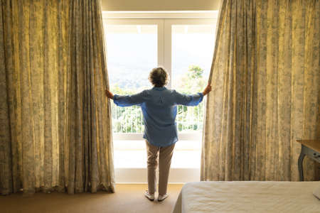 Senior caucasian woman standing next to window and widening courtains in bedroom. retreat, retirement and senior lifestyle concept.の写真素材