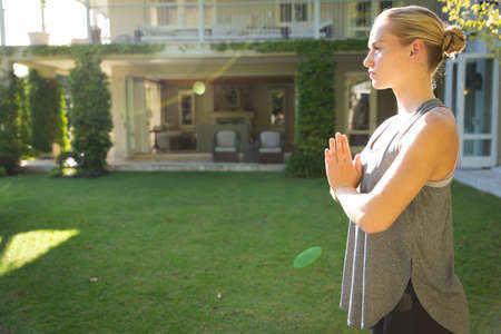 Tranquil caucasian woman practicing yoga in sunny garden, standing with hands pressed together. health, fitness and wellbeing, spending quality time at home.の写真素材