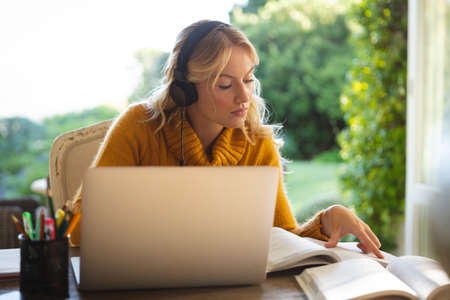 Caucasian woman working in living room at home, wearing headphones and using laptop, reading book. flexible working from home with technology.の写真素材