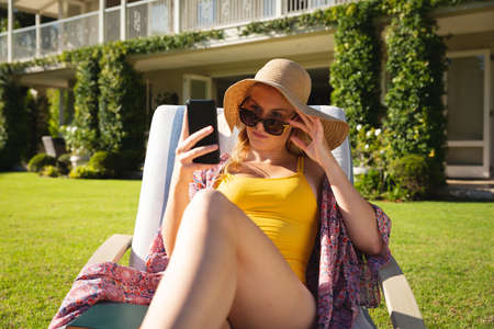 Caucasian woman sitting in sunny garden wearing sunhat and sunglasses using smartphone. spending free time at home with technology.の写真素材
