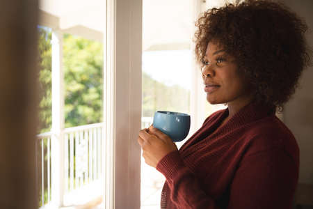 Happy african american woman looking out of sunny window, holding coffee cup and smiling. spending...