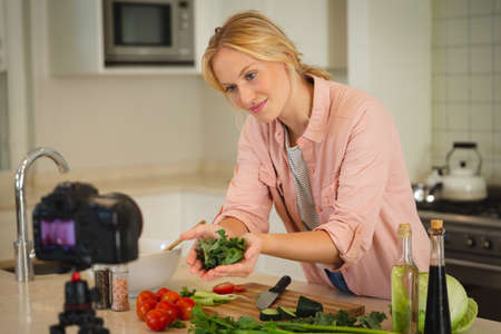 Smiling caucasian woman in kitchen holding vegetables and looking at camera, making cooking vlog....