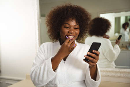 Smiling african american woman in bathroom wearing bathrobe, brushing teeth and using smartphone....