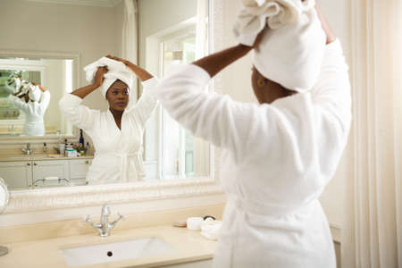 African american woman in bathroom wearing bathrobe, looking in mirror and putting towel on head. health, beauty and wellbeing, spending quality time at home.の写真素材