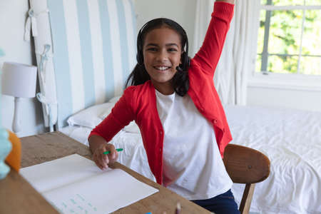 Smiling african american girl having video call lesson wearing headphones and raising hand at home....