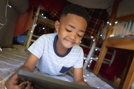 Happy african american boy at home, lying in blanket fort and using tablet. childhood with technology, spending free time at home.の写真素材