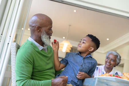 Happy african american grandson and grandparents sitting at table talking during breakfast. family...