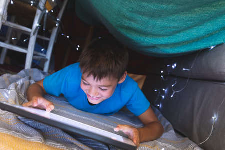 Caucasian boy smiling, lying in blanket fort using tablet at home. childhood with technology,...