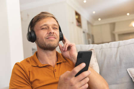 Caucasian man sitting on couch, wearing headphones and using smartphone at home. enjoying free time...