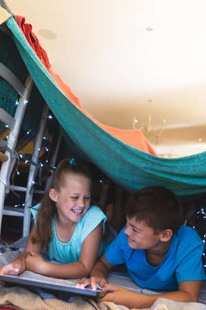 Caucasian brother and sister smiling, lying in blanket fort using tablet at home. childhood with technology, spending free time at home.