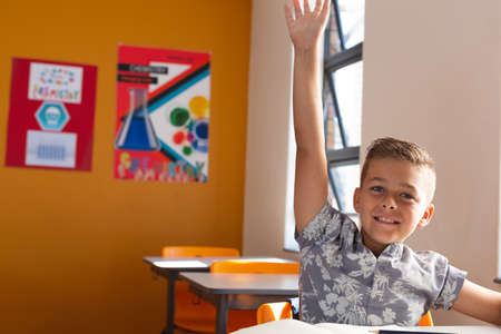 Happy caucasian schoolboy sitting at desk in classroom raising hand during lesson. childhood and...