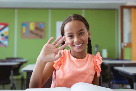 Portrait of smiling mixed race schoolgirl sitting at desk in classroom waving hand. childhood and education at elementary school.の写真素材