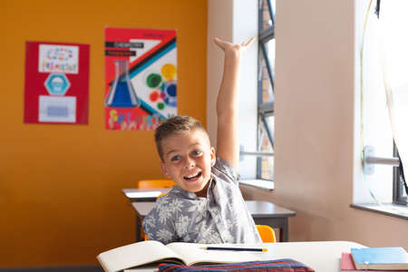 Excited caucasian schoolboy sitting at desk in classroom raising hand during lesson. childhood and education at elementary school.の写真素材