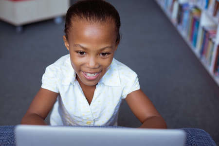 Smiling african american schoolgirl at desk in school library using laptop. childhood, technology and education at elementary school.の写真素材