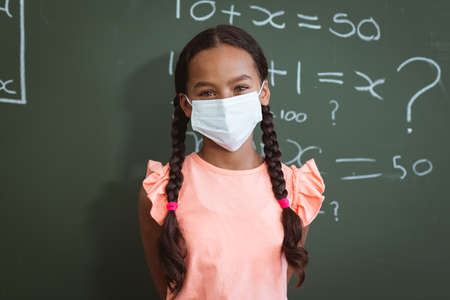 Portrait of mixed race schoolgirl in face mask standing in front of chalkboard in maths classroom. childhood and education at elementary school during  pandemic. .の写真素材