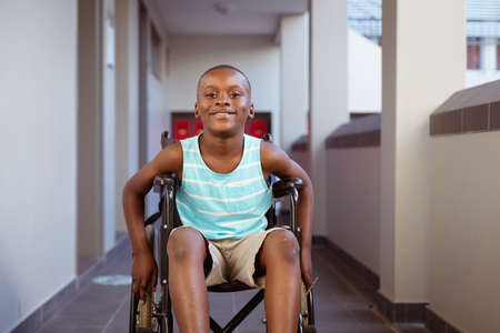 Portrait of smiling disabled african american schoolboy sitting in wheelchair in school corridor. childhood and education at elementary school.の写真素材