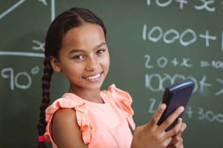 Portrait of smiling mixed race schoolgirl standing at chalkboard in classroom using smartphone. childhood, technology and education at elementary school.の写真素材