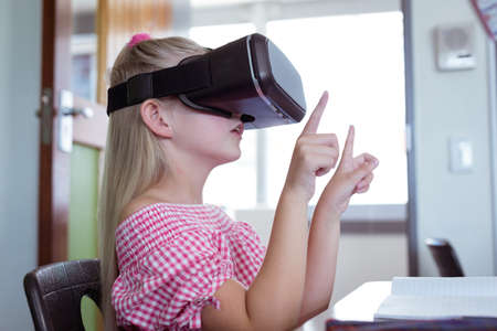 Caucasian schoolgirl sitting at desk in classroom wearing vr headset. childhood, technology and education at elementary school.の写真素材
