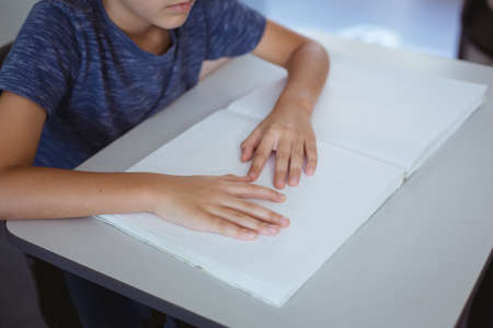 Blind caucasian schoolboy sitting at desk reading braille book with fingers. childhood and education at elementary school.の写真素材