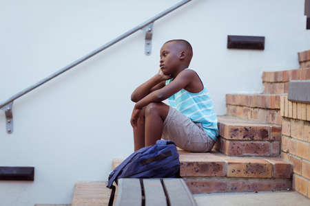 Bored african american schoolboy sitting on steps in school yard. childhood and education at elementary school.の写真素材