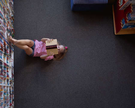 Overhead view of caucasian schoolgirl lying on floor reading book in school library. childhood and...