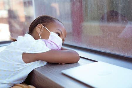 African american schoolgirl wearing face mask sitting in school classroom looking out of window. childhood and education at elementary school during  pandemic. .の写真素材