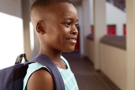 Smiling african american schoolboy wearing schoolbag standing in school corridor. childhood and...