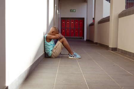 Upset african american schoolboy sitting on floor in school corridor with head in hands. childhood...
