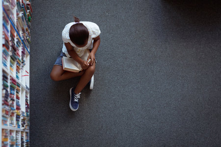 Overhead view of african american schoolgirl sitting on floor reading book in school library. childhood and education at elementary school.の写真素材