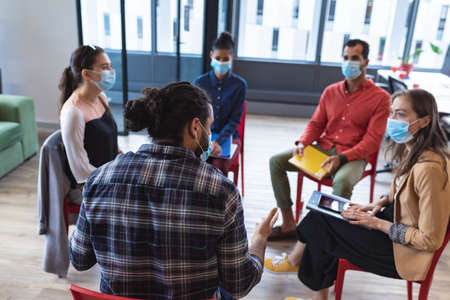 Diverse group of creative colleagues wearing face masks talking at meeting. work at a modern office during covid 19 coronavirus pandemic.の写真素材