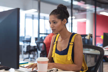 Mixed race female creative worker sitting at desk using computer. modern office of a creative design business.の写真素材