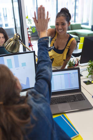 Two diverse female creative colleagues sitting at desk high-fiving and smiling. modern office of a creative design business.の写真素材