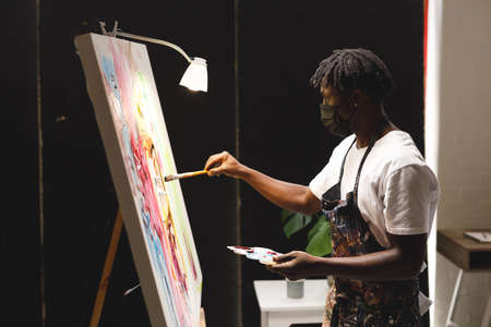African american male painter wearing face mask painting on canvas in art studio. creation and...