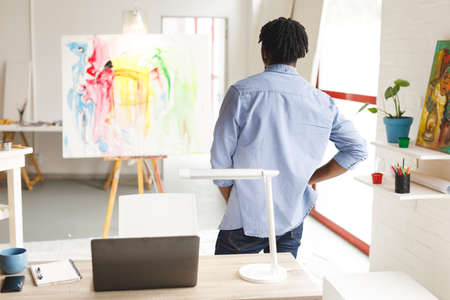 African american male painter at work looking at artwork in art studio. creation and inspiration at...