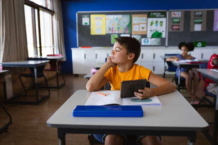 Caucasian boy holding digital tablet while sitting on the desk in the class at elementary school. school and education conceptの写真素材