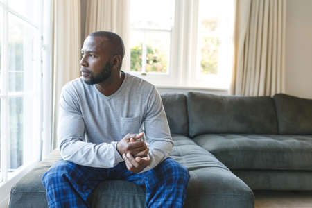 Thoughtful african american man sitting on couch looking out of window in living room. spending time alone at home.の写真素材