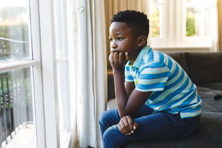 Thoughtful african american boy sitting and looking out of window in living room. spending time alone at home.の写真素材