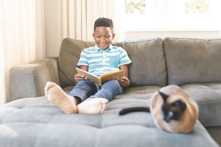 Smiling african american boy reading book and sitting on couch with cat in living room. spending time alone at home.の写真素材