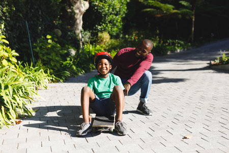 Portrait of african american father with son smiling and playing with skateboard in garden. family spending time at home.の写真素材