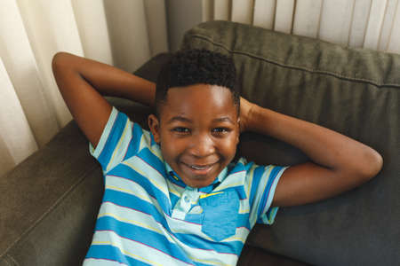 Portrait of smiling african american boy lying back on couch in living room. spending time alone at home.の写真素材