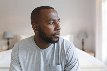 Thoughtful african american man sitting on bed and looking away in bedroom. spending time alone at...