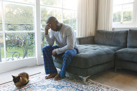 Worried, sad african american man thinking, sitting on couch holding head in living room with cat....